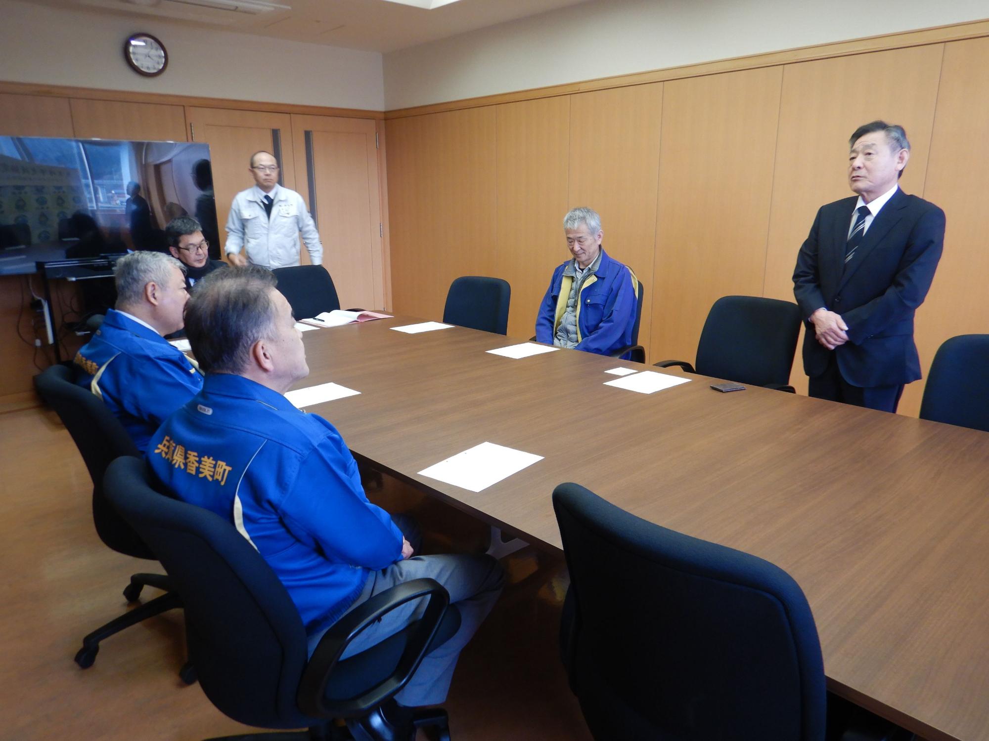 会議室のような場所で黒いスーツの男性が青いジャケットの男性らに挨拶している写真