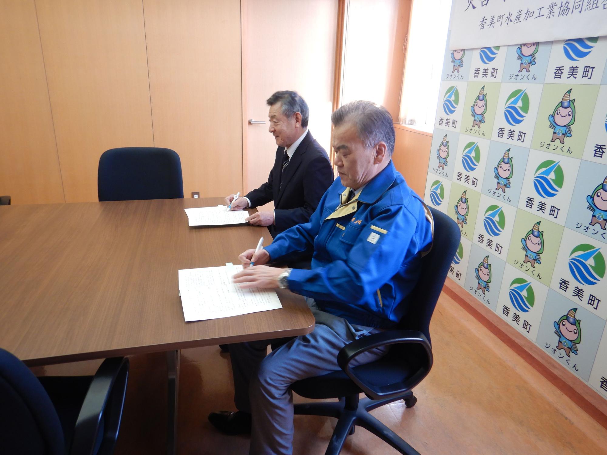 机の上の書類に目を通す青いジャケットの男性とその奥に黒いスーツの男性が隣に座っている写真