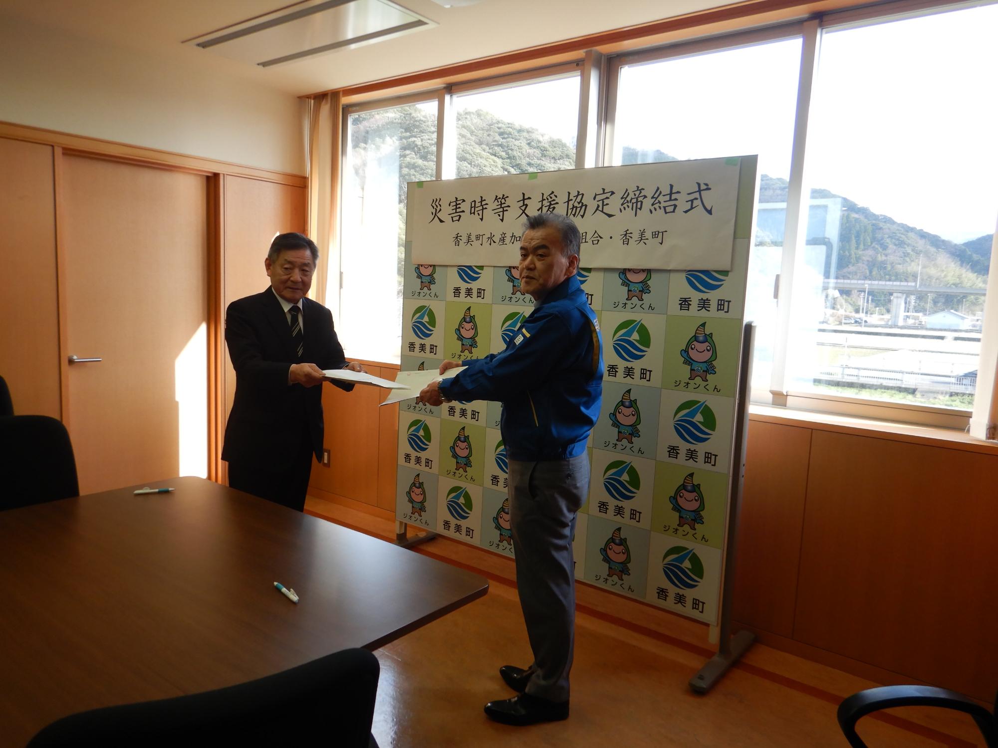 窓際で2人の男性が向かい合って書類を交換している写真