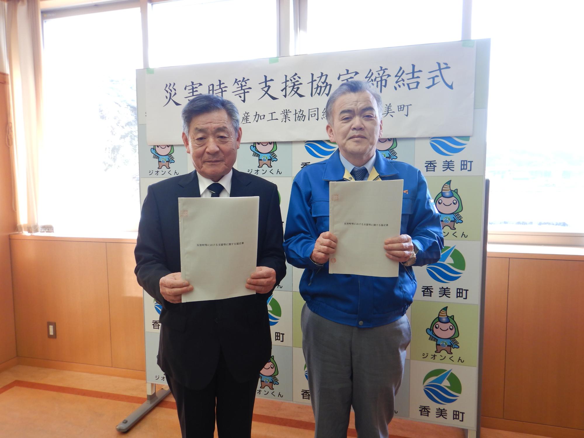 黒いスーツの男性と青いジャケットの男性が書類を両手に持って一緒に写っている写真