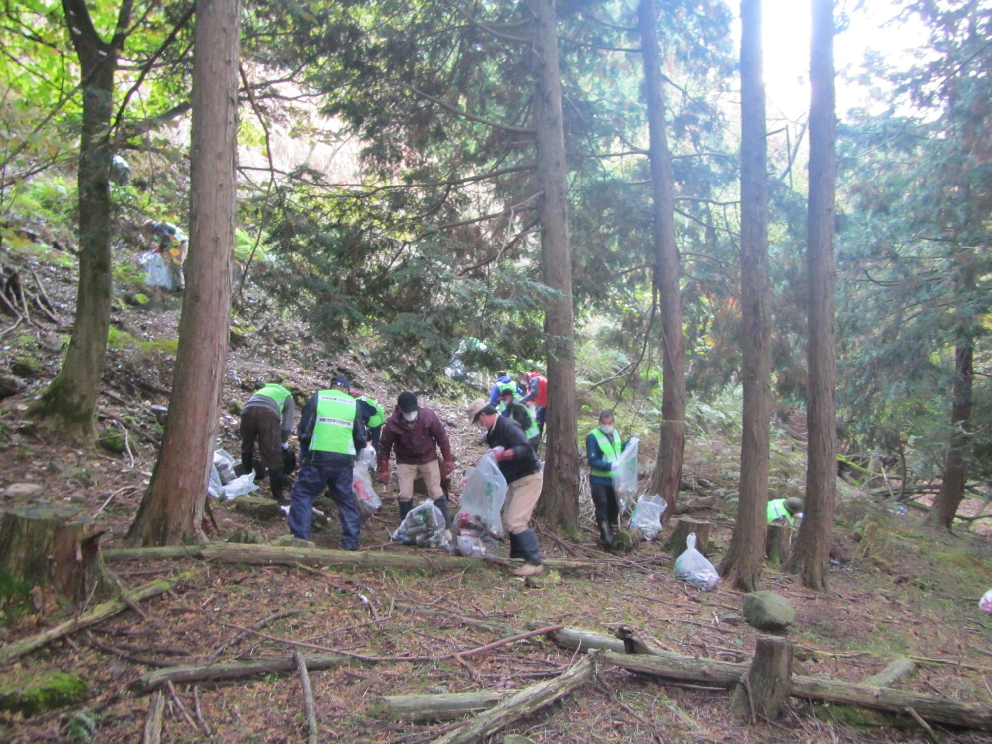 山の茂みで、作業服や帽子を身につけた人たちがゴミ拾いをしている写真