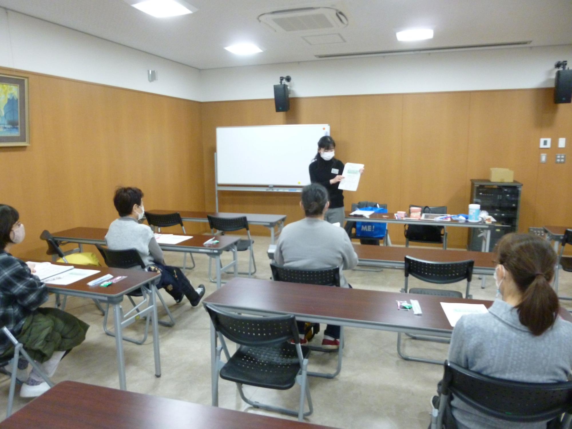 会議室で女性が書面を見せながら話しているのを複数人が聞いている写真